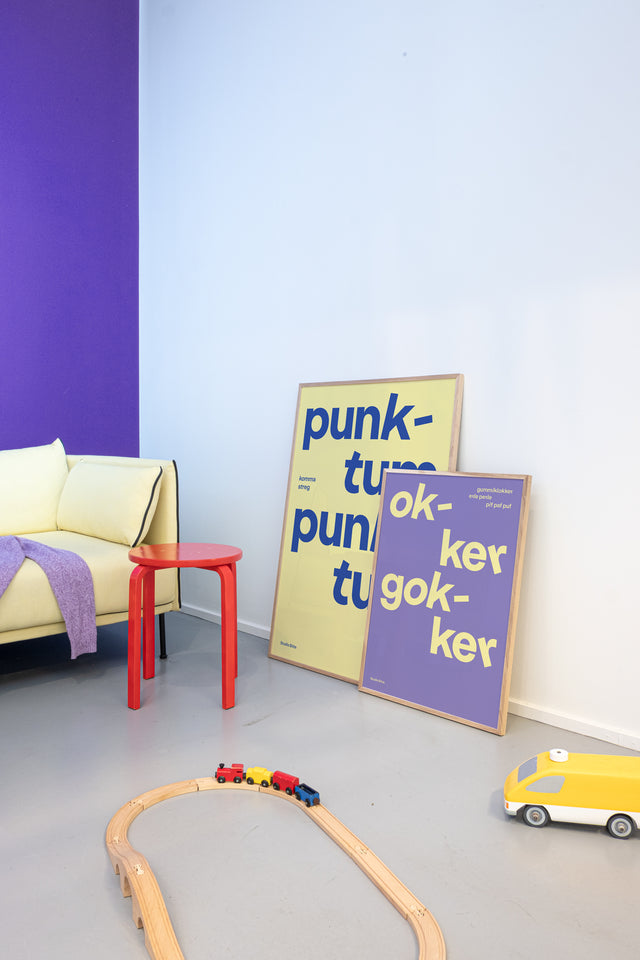 Gul "punktum punktum" børneplakat og lilla "okker gokker" børneplakat på børneværelse med gul sofa og lilla farvevæg
